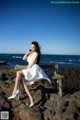 A woman in a white dress sitting on a bench by the ocean.