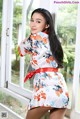 A woman in a white and orange floral dress posing by a window.