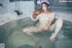 A naked woman sitting in a hot tub holding a cup.