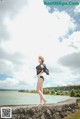 A woman in a black bikini standing on a stone wall by the water.