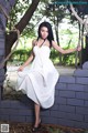 A woman in a white dress leaning against a brick wall.